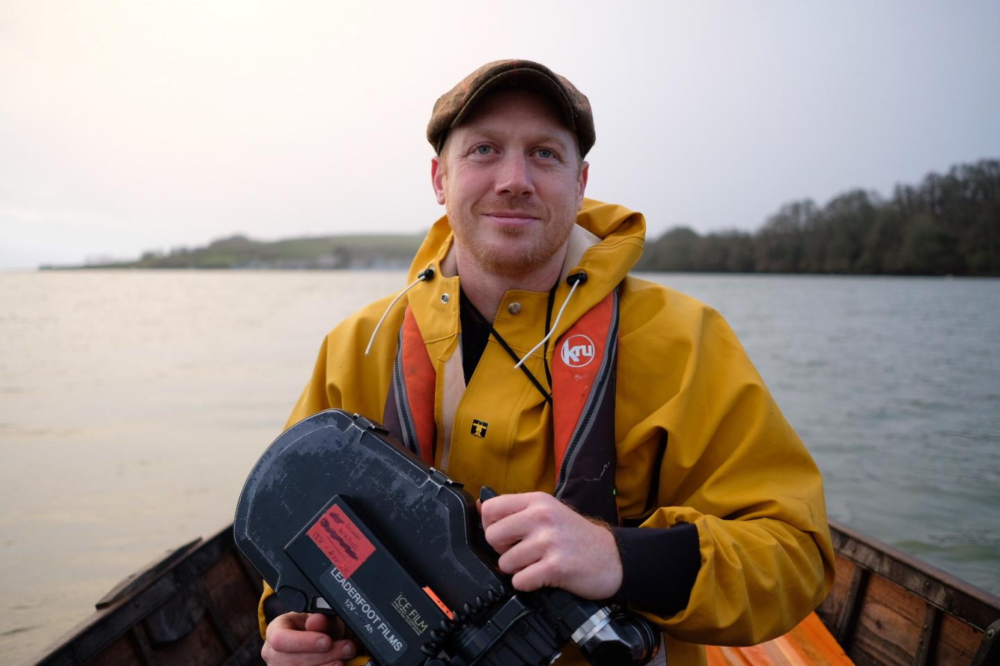 A man, filmmaker Huw Wahl, holding a camera, wearing a yellow waterproof coat and a life jacket and a brown flat cap.