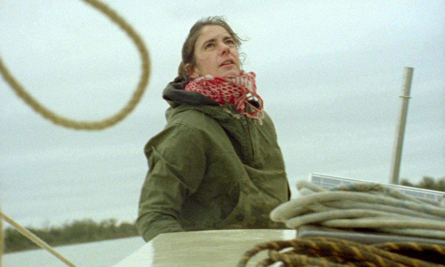 A woman, Rose Ravetz, standing on a boat wearing a green anorak and red scarf.