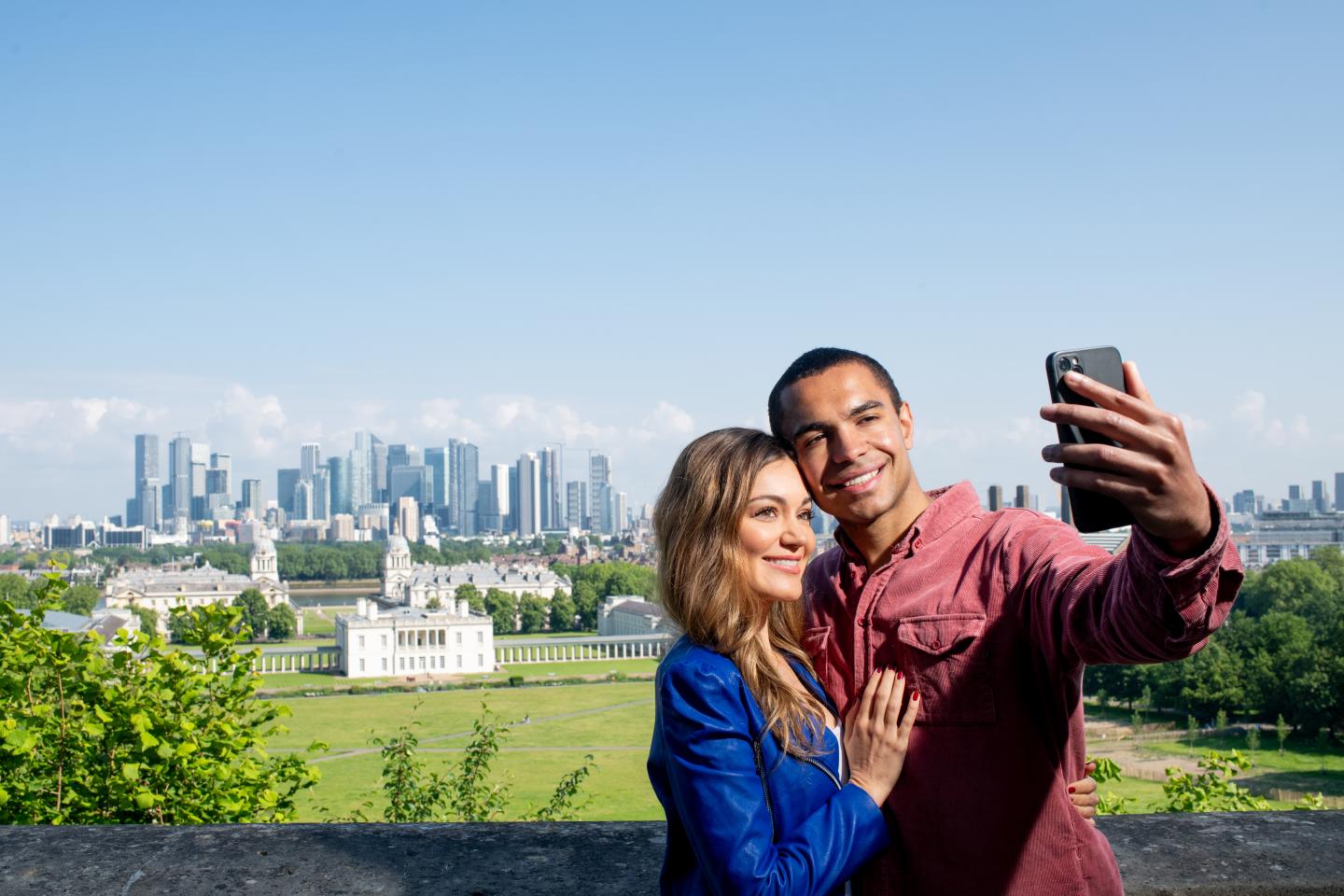 A man and a woman take a selfie at the top of Greenwich Park, with the River Thames and Canary Wharf in the background