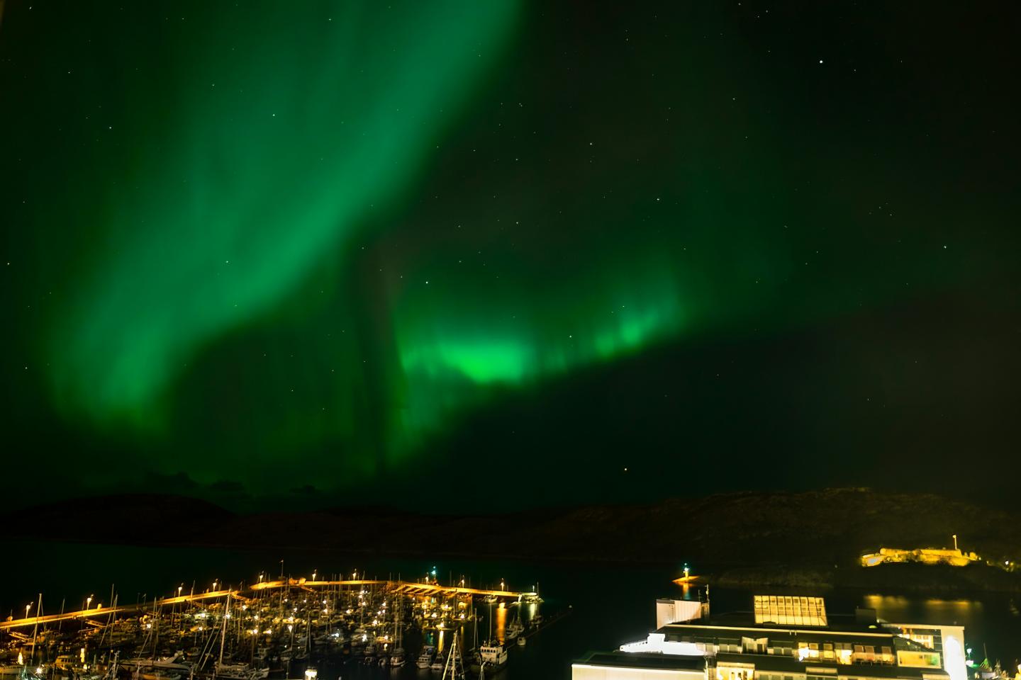 A photo of green aurora in the night time sky with brightly lit boats beneath