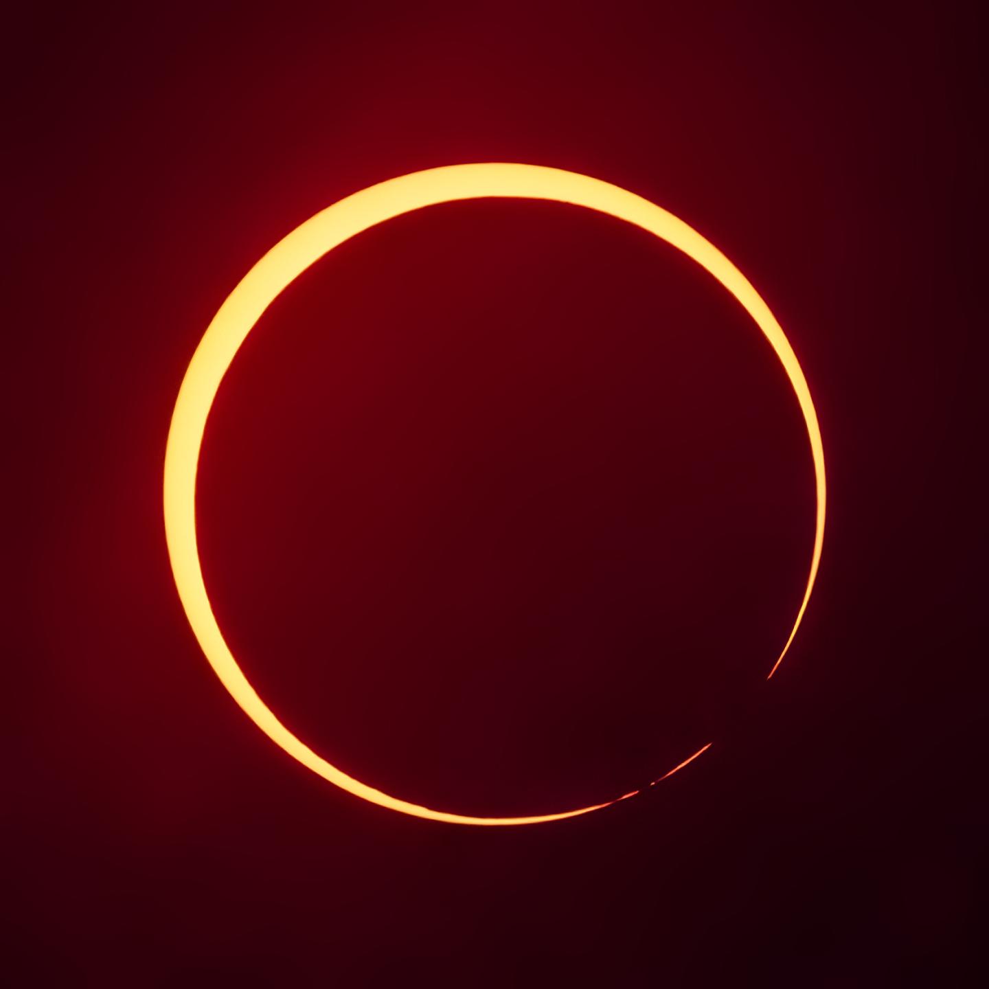 Square image of dark red/black background with an annular solar eclipse taking place. The Sun is a round yellow disk, and the Moon is in front of it as a black/red disk, but the Moon is not centrally aligned with the sun and is touching the south east side. Parts of the sun where the moon is touching the edge are obscured by shadows of lunar mountains.