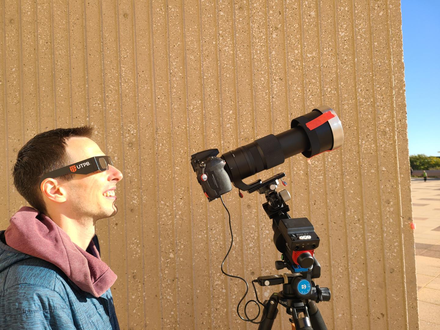 A man standing in direct sunlight wearing eclipse glasses and looking up towards the Sun. There is a camera in front of him which has a very long lens and a solar filter taped over the end, it is hooked up with wires to a complex tracking mount and tripod