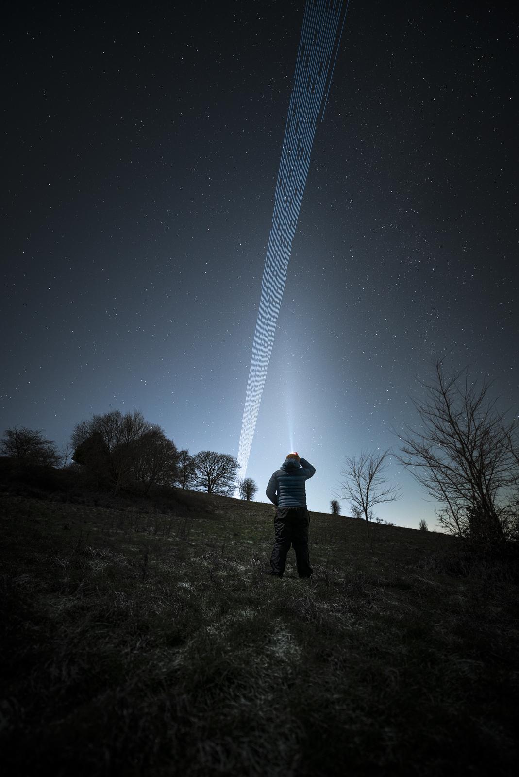 Image from further down a hill looking up at a man standing with a head torch on pointing at the sky. A few trees dot the landscape, while in the sky there are many stars but a long trail of wavy, dotted lines in a straight line, which are satellite trails.