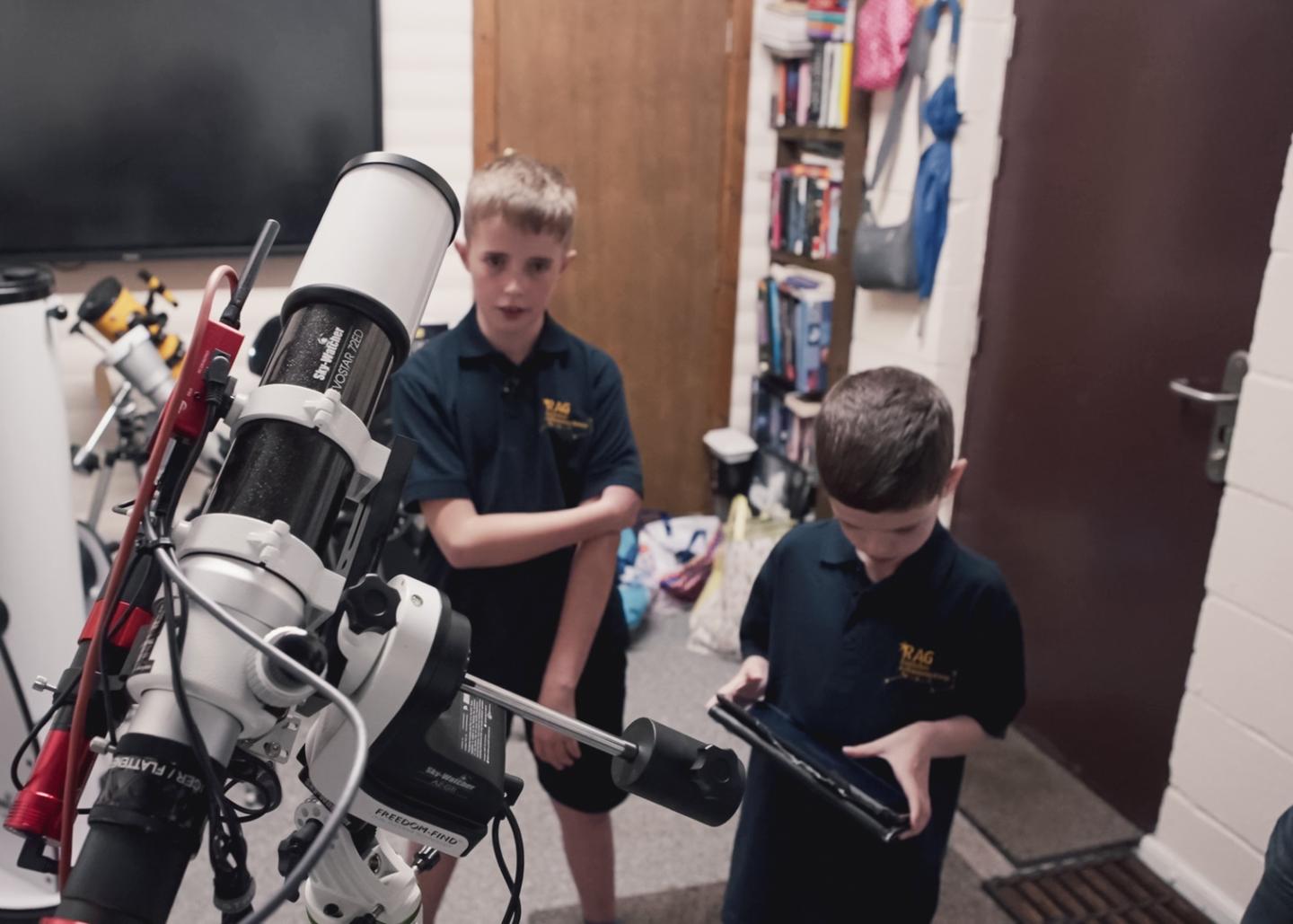 Two young boys operate a telescope. One is holding an iPad, while the other looks up at the scope