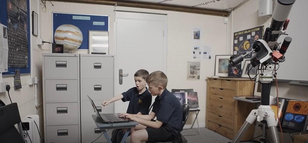 Photo of two children looking at a computer screen, the younger brother is looking happy and surprised and pointing at the screen. They are sitting inside a room decorated with astronomy objects, including a model of jupiter and a framed astronomy photograph.