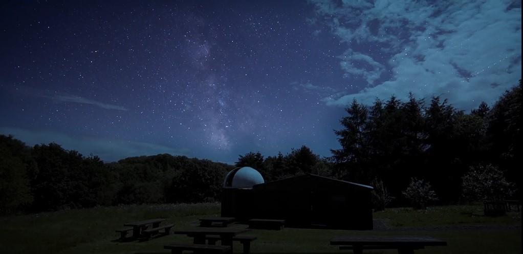 Night photo of small observatory surrounded by trees, in the sky above is the Milky Way against a dark blue sky