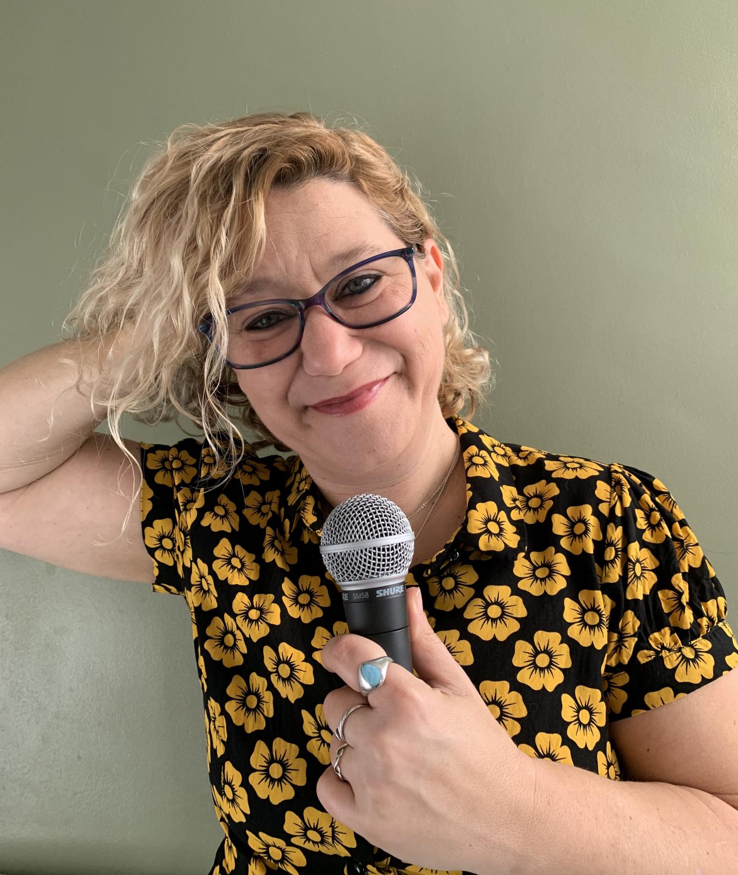 Photo of Leslie Gold against a mint green background, with one hand on their head and the other holding a microphone