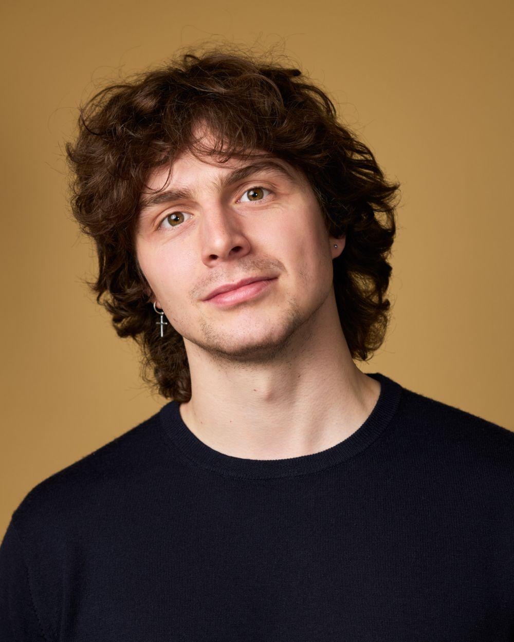 Headshot of Sam Williams in front of a yellow background wearing a black t shirt, looking and slightly smiling at the camera