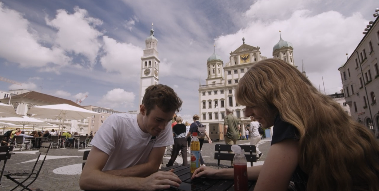 Image of two people sitting in a historic town square opposite each other, looking down at a phone and smiling