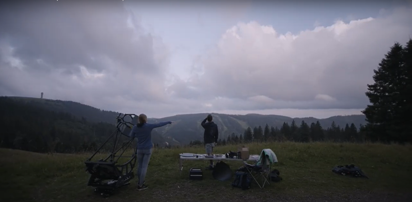 Photo of two people standing at the top of a small mountain in an alpine region, one is pointing at the sky which is cloudy, and one is looking up at the same place next to a table which has photography equipment on it