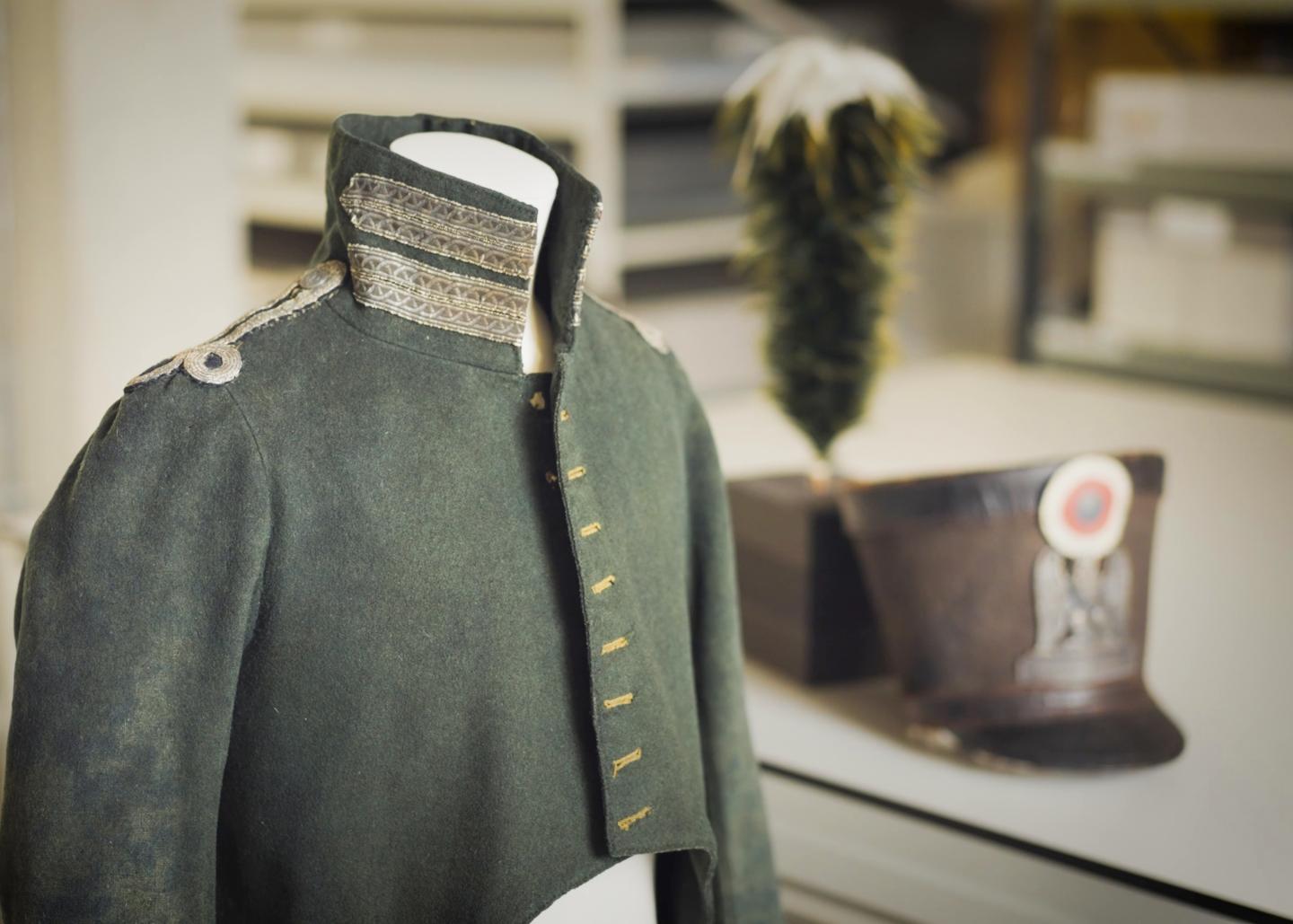 A well-worn, dark green military uniform with silver and yellow detailing. The uniform is on a mannequin, and on the table behind it is a large hat and plume of feathers