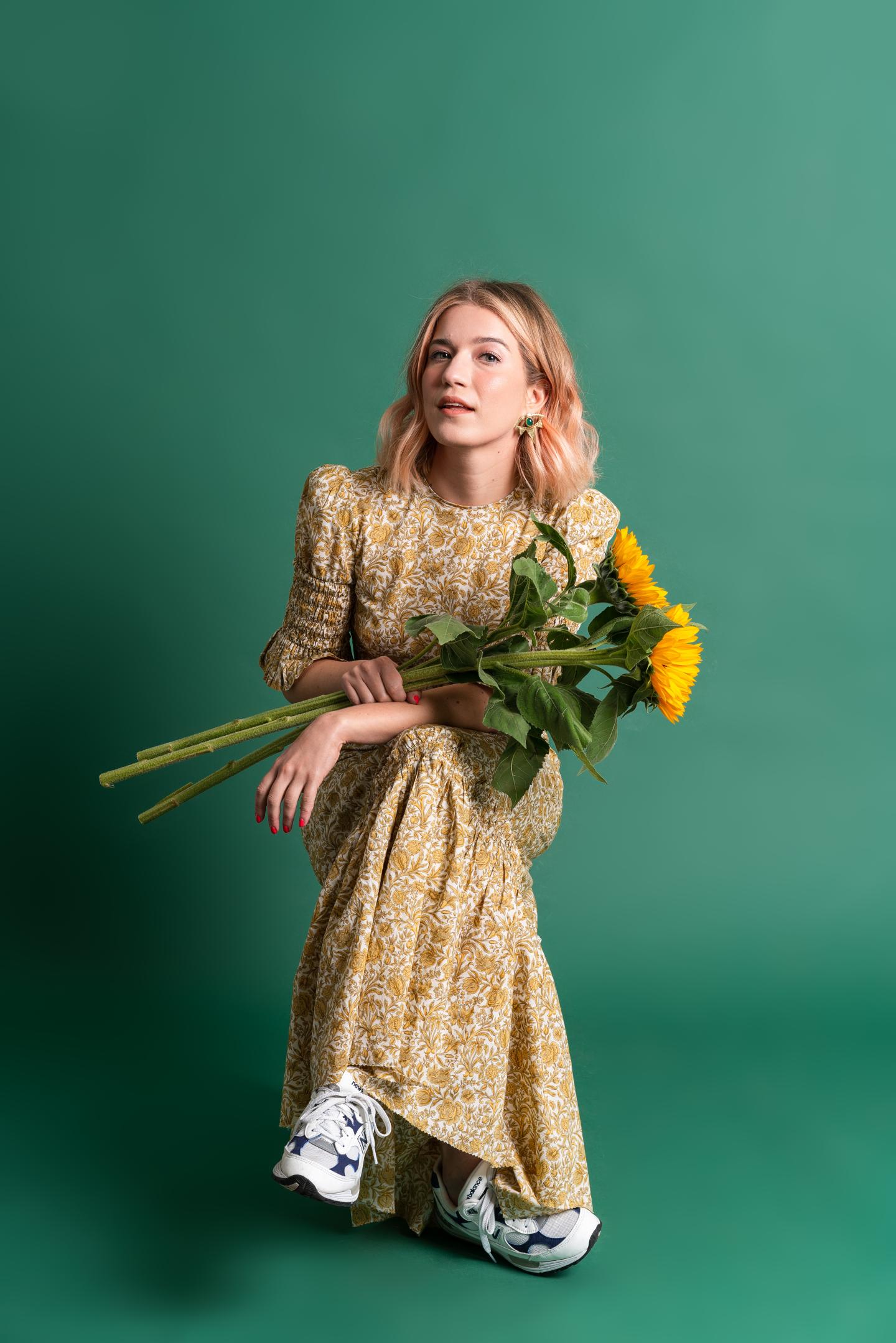 Photo of a woman sitting in front of a green background holding sunflowers and wearing a yellow sundress with trainers