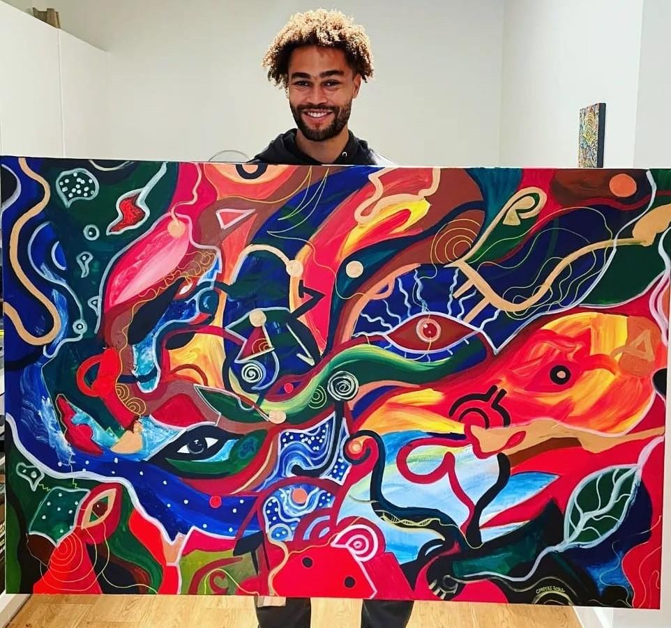 Artist Joel Sydenham smiling with a large, brightly coloured, abstract painting.