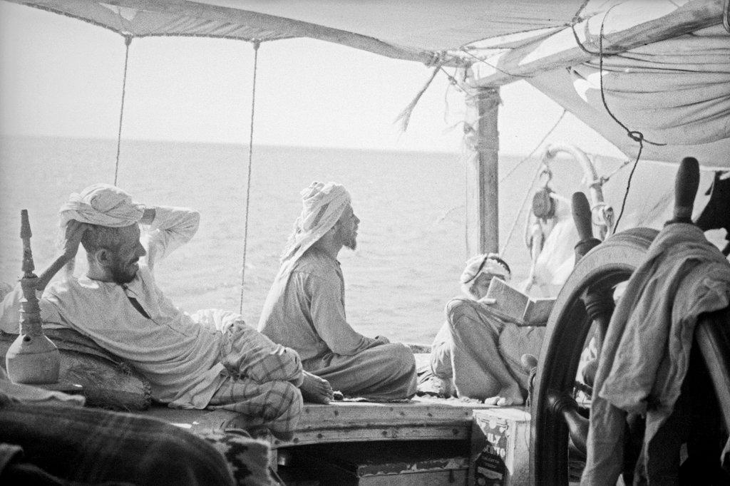 Black and white photo of men wearing head coverings lounging on a covered deck of a ship at sea, with the ship's wheel in the foreground