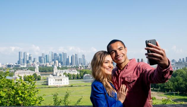 A man and a woman take a selfie at the top of Greenwich Park, with the River Thames and Canary Wharf in the background